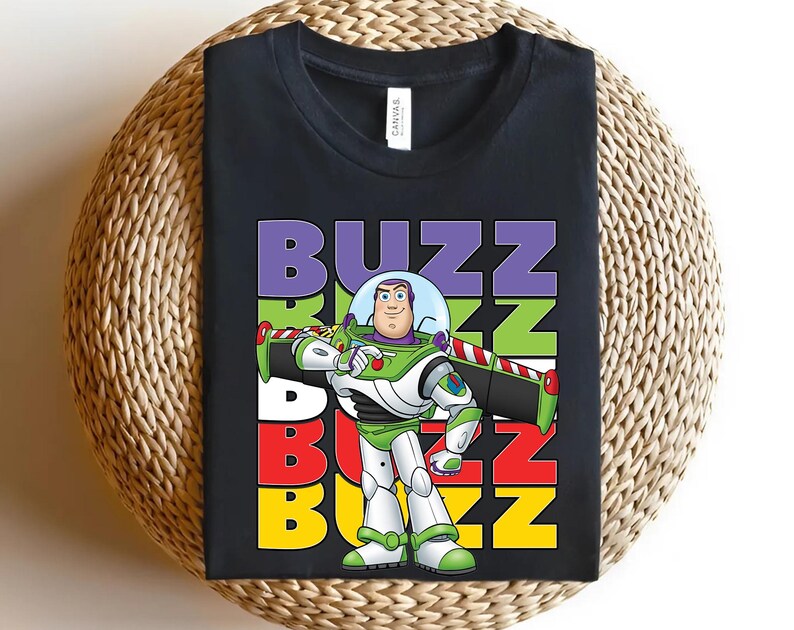 Puede incluir: Camiseta negra con un gr&aacute;fico de Buzz Lightyear de Toy Story. El dise&ntilde;o incluye la palabra "BUZZ" en letras apiladas de colores, con Buzz Lightyear en su traje espacial verde, blanco y morado. La camiseta se muestra sobre una superficie tejida.