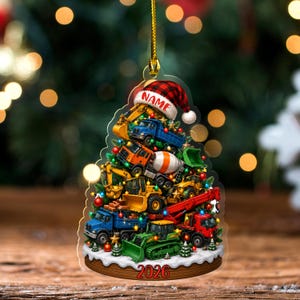 May include: A Christmas tree-shaped ornament featuring a stack of construction vehicles including dump trucks, excavators, and a cement mixer. A red and black plaid Santa hat with the text NAME sits on top, and the base displays the year 2026.