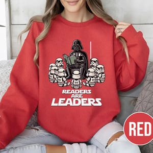 K&ouml;nnte beinhalten: Rotes Sweatshirt mit einer Grafik von Darth Vader, der Stormtroopern vorliest. Der Text "READERS ARE LEADERS" ist darunter aufgedruckt. Das Sweatshirt hat eine einfarbige rote Farbe. Das Design ist eine spielerische Interpretation eines beliebten Science-Fiction-Themas.