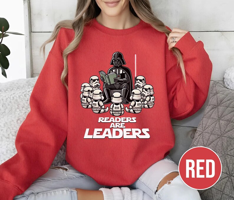 K&ouml;nnte beinhalten: Rotes Sweatshirt mit einer Grafik von Darth Vader, der Stormtroopern vorliest. Der Text "READERS ARE LEADERS" ist darunter aufgedruckt. Das Sweatshirt hat eine einfarbige rote Farbe. Das Design ist eine spielerische Interpretation eines beliebten Science-Fiction-Themas.