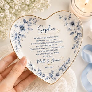 May include: A heart-shaped ceramic trinket dish with a gold-trimmed edge, featuring blue floral illustrations and printed text: Sophia, We did not get to choose you, that honor was our sons, But if we could have picked his match, you still would be the one.