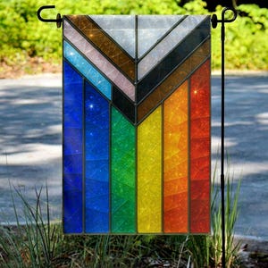 May include: A rectangular garden flag with a stained glass design. The flag features a vibrant rainbow of colors, including blue, green, yellow, orange, and red. The top portion has a chevron pattern with brown, black, and white sections. The flag is hanging on a black metal stand.