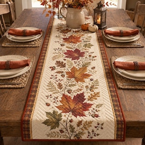 May include: A quilted table runner featuring a central pattern of autumn leaves, acorns, and berries in shades of red, orange, and brown. The runner has a cream-colored background with a dark plaid border and is displayed on a wooden dining table.