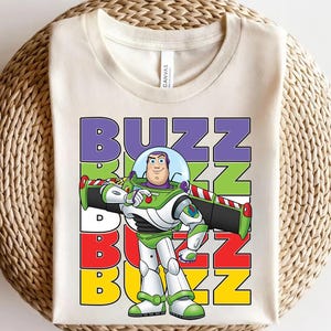 Puede incluir: Camiseta color crema con un gr&aacute;fico de Buzz Lightyear de Toy Story. El dise&ntilde;o incluye la palabra "BUZZ" en letras apiladas y coloridas, con Buzz Lightyear en su traje espacial verde, blanco y morado caracter&iacute;stico.