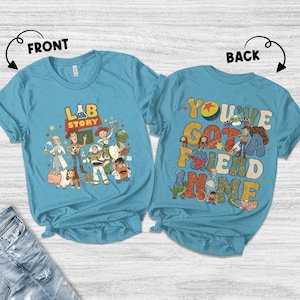 Op de afbeelding: Een lichtblauw T-shirt met een kleurrijke afbeelding van Toy Story-personages. De voorkant van het shirt zegt "LAB STORY" en de achterkant zegt "YOU'VE GOT A FRIEND IN ME".