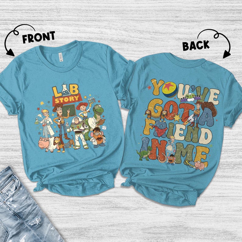 Op de afbeelding: Een lichtblauw T-shirt met een kleurrijke afbeelding van Toy Story-personages. De voorkant van het shirt zegt "LAB STORY" en de achterkant zegt "YOU'VE GOT A FRIEND IN ME".