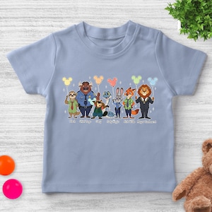 May include: A light blue toddler t-shirt featuring a colorful graphic of characters from the movie Zootopia. The characters include Flash, Chief Bogo, Gary, Judy Hopps, Nick Wilde, and Mayor Lionheart. Each character is holding a balloon.