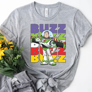 Puede incluir: Una camiseta gris con un gr&aacute;fico de Buzz Lightyear de Toy Story. La palabra "BUZZ" est&aacute; apilada verticalmente en morado, verde, rojo y amarillo. Buzz Lightyear se muestra con su traje espacial verde y blanco.