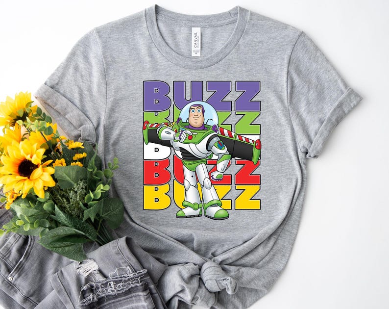 Puede incluir: Una camiseta gris con un gr&aacute;fico de Buzz Lightyear de Toy Story. La palabra "BUZZ" est&aacute; apilada verticalmente en morado, verde, rojo y amarillo. Buzz Lightyear se muestra con su traje espacial verde y blanco.