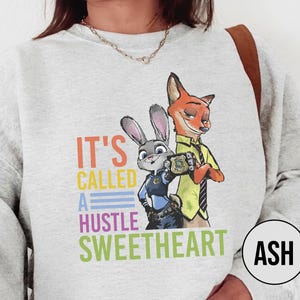 Puede incluir: Una sudadera gris claro con un gr&aacute;fico colorido de Judy Hopps y Nick Wilde de Zootopia, con el texto "IT'S CALLED A HUSTLE SWEETHEART". El dise&ntilde;o incluye a los personajes con sus uniformes de polic&iacute;a.
