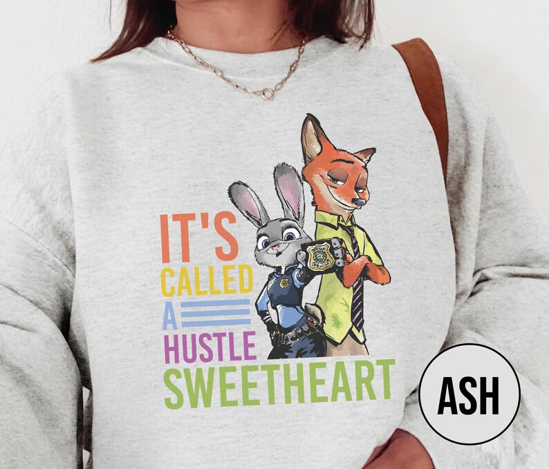 Puede incluir: Una sudadera gris claro con un gr&aacute;fico colorido de Judy Hopps y Nick Wilde de Zootopia, con el texto "IT'S CALLED A HUSTLE SWEETHEART". El dise&ntilde;o incluye a los personajes con sus uniformes de polic&iacute;a.