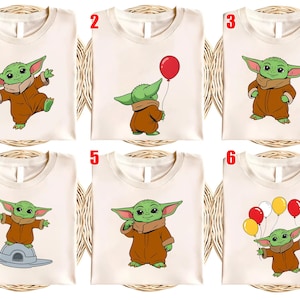 May include: Cream-colored t-shirts featuring cartoon illustrations of a green alien character in a brown outfit. The designs include the character holding a red balloon, standing on a spaceship, and holding multiple balloons. The shirts are displayed on a woven background.