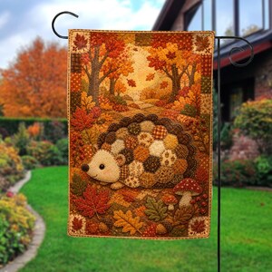 May include: A rectangular garden flag featuring a patchwork-style hedgehog with a brown and orange patterned body. The scene includes autumn leaves, acorns, and mushrooms against a backdrop of a forest path with golden trees and falling foliage.
