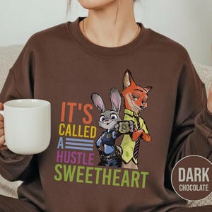 Puede incluir: Sudadera marr&oacute;n chocolate oscuro con un gr&aacute;fico de Judy Hopps y Nick Wilde de Zootopia. El texto "IT'S CALLED A HUSTLE SWEETHEART" est&aacute; impreso en letras coloridas. Una taza de caf&eacute; blanca se sostiene en la mano izquierda.