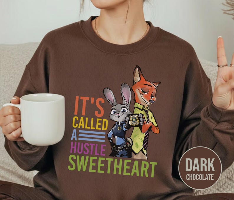 Puede incluir: Sudadera marr&oacute;n chocolate oscuro con un gr&aacute;fico de Judy Hopps y Nick Wilde de Zootopia. El texto "IT'S CALLED A HUSTLE SWEETHEART" est&aacute; impreso en letras coloridas. Una taza de caf&eacute; blanca se sostiene en la mano izquierda.