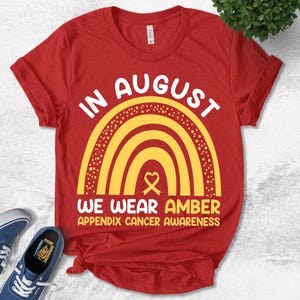 May include: A red short-sleeved t-shirt featuring a yellow rainbow graphic with a heart-shaped ribbon. The text reads: IN AUGUST WE WEAR AMBER APPENDIX CANCER AWARENESS. The design is centered on the chest of the shirt, which has a crew neckline.