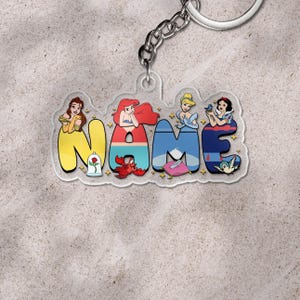 May include: A clear acrylic keychain with the word "NAME" in colorful bubble letters. Each letter features a Disney princess: Belle, Ariel, Cinderella, and Snow White. The keychain includes a silver chain and keyring.
