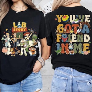 Op de afbeelding: Zwarte t-shirts met kleurrijke afbeeldingen. E&eacute;n shirt heeft de tekst "LAB STORY" met cartoonfiguren. Het andere shirt toont "YOU'VE GOT A FRIEND IN ME" met meer cartoonfiguren. Beide shirts hebben een vintage stijl.