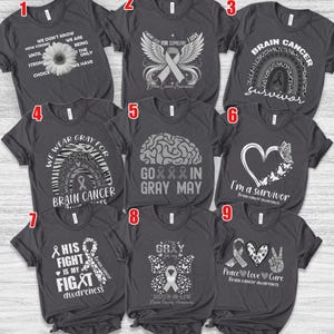 May include: Nine dark grey t-shirts, each featuring a different white graphic and text design related to brain cancer awareness. Designs include ribbons, hearts, and inspirational quotes. The t-shirts are short-sleeved.