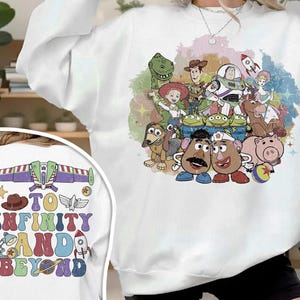 May include: White sweatshirt featuring a colorful graphic of Toy Story characters. The design includes Woody, Buzz Lightyear, and other characters. The back of the sweatshirt has the text "TO INFINITY AND BEYOND" with a rocket ship and other graphics.