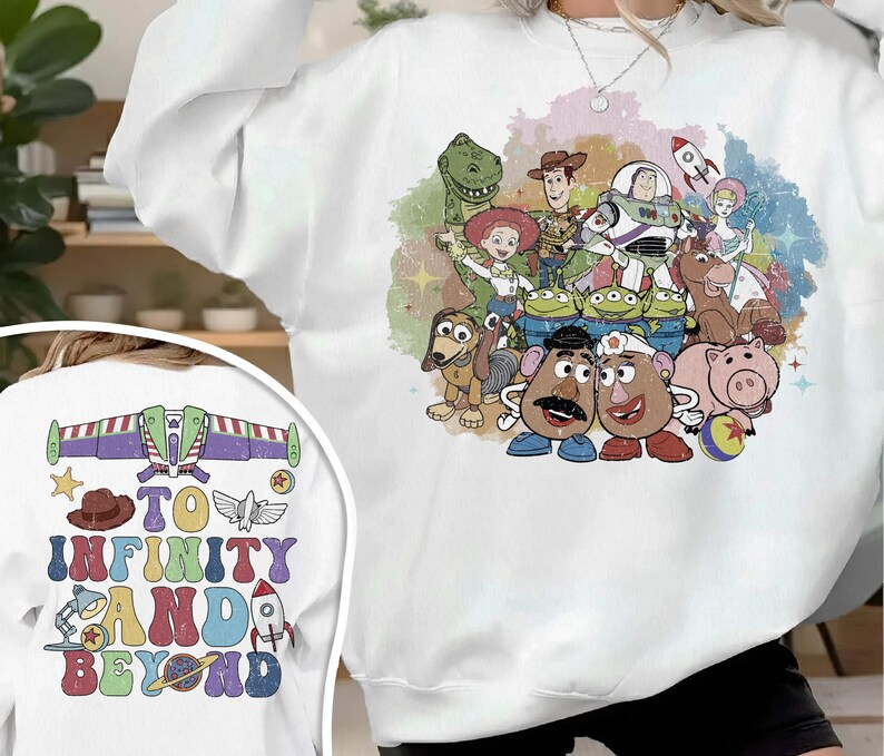 May include: White sweatshirt featuring a colorful graphic of Toy Story characters. The design includes Woody, Buzz Lightyear, and other characters. The back of the sweatshirt has the text "TO INFINITY AND BEYOND" with a rocket ship and other graphics.