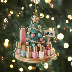 May include: A Christmas tree ornament featuring a teal Christmas tree with pink, blue, and gold ornaments. The ornament includes a sign that reads "SARAH", nail polish bottles, and a small gift box. The ornament is suspended by a gold string.