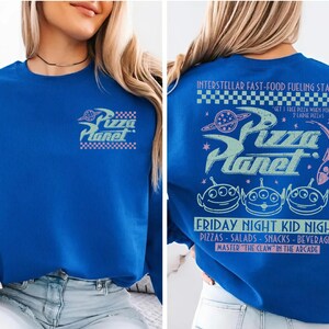 May include: Royal blue sweatshirt featuring the Pizza Planet logo on the front and back. The back design includes the text "Interstellar Fast-Food Fueling Station" and "Friday Night Kid Night". The design also features three green alien characters.