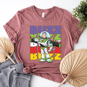 Puede incluir: Una camiseta de color malva con un gr&aacute;fico de Buzz Lightyear de Toy Story. El dise&ntilde;o incluye la palabra "BUZZ" apilada verticalmente en morado, verde, blanco, rojo y amarillo. Buzz Lightyear est&aacute; representado en su traje espacial verde y blanco.
