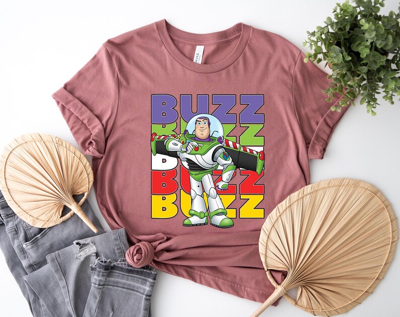 Puede incluir: Una camiseta de color malva con un gr&aacute;fico de Buzz Lightyear de Toy Story. El dise&ntilde;o incluye la palabra "BUZZ" apilada verticalmente en morado, verde, blanco, rojo y amarillo. Buzz Lightyear est&aacute; representado en su traje espacial verde y blanco.