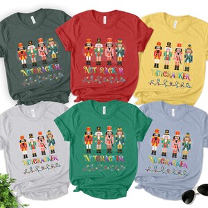 May include: Six short-sleeved crew neck t-shirts in dark green, red, yellow, light gray, green, and light blue. Each shirt features a graphic of four nutcracker figures above the multicolored text NUTCRACKER and a string of colorful Christmas lights.