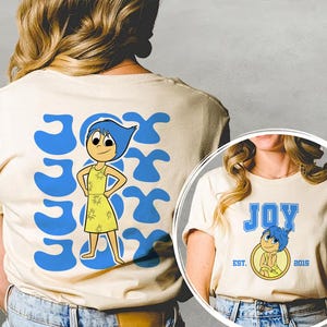 May include: Cream-colored t-shirt featuring a cartoon character with blue hair and a yellow dress. The word "JOY" is printed in blue, along with the character's image. The back of the shirt has the word "JOY" repeated in a blue design.