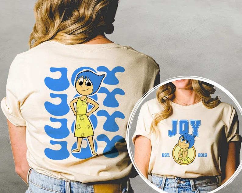 May include: Cream-colored t-shirt featuring a cartoon character with blue hair and a yellow dress. The word "JOY" is printed in blue, along with the character's image. The back of the shirt has the word "JOY" repeated in a blue design.
