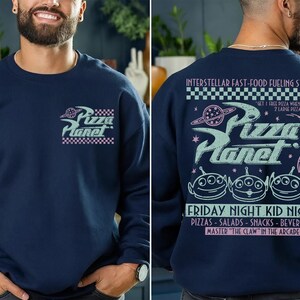 May include: Navy blue sweatshirt featuring the Pizza Planet logo on the front and back. The front logo is a stylized design with the words "Pizza Planet" and a checkered border. The back of the sweatshirt has the text "INTERSTELLAR FAST-FOOD FUELING STATION".