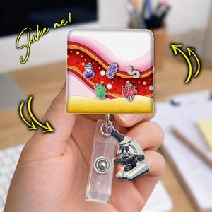 May include: A square badge reel featuring a blood vessel illustration with floating cartoon microbes, including a purple potion, a green bacterium, a pink cell, and a worm. A small microscope charm hangs from the clear plastic strap below the reel.
