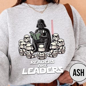 K&ouml;nnte beinhalten: Hellgraues Sweatshirt mit einer Grafik von Darth Vader, der Stormtroopern vorliest. Der Text "Readers Are Leaders" ist unter dem Bild aufgedruckt. Ein rotes Lichtschwert ist ebenfalls zu sehen.