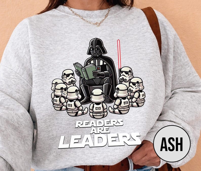 K&ouml;nnte beinhalten: Hellgraues Sweatshirt mit einer Grafik von Darth Vader, der Stormtroopern vorliest. Der Text "Readers Are Leaders" ist unter dem Bild aufgedruckt. Ein rotes Lichtschwert ist ebenfalls zu sehen.