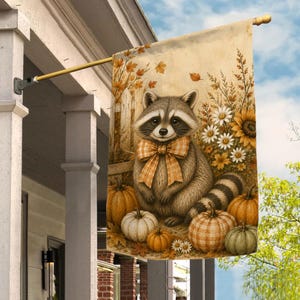 May include: A rectangular garden flag featuring a central illustration of a raccoon wearing a plaid bow tie, surrounded by orange and white pumpkins, sunflowers, and daisies against a light beige background with autumn leaves and a wooden fence.