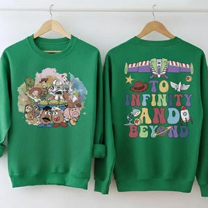 May include: Two green sweatshirts. One features a colorful graphic of Toy Story characters. The other has a Buzz Lightyear design with the phrase "To Infinity and Beyond" in a retro font. Both are displayed on wooden hangers.