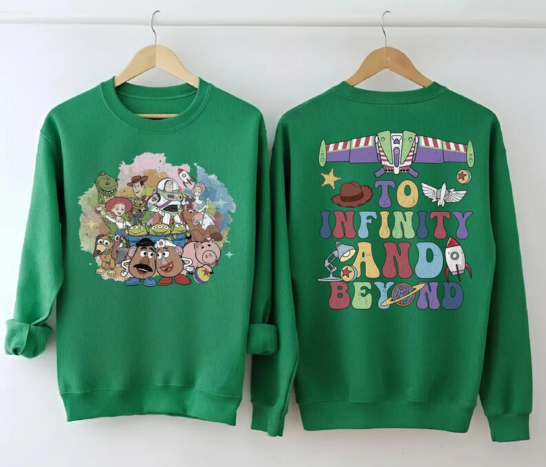 May include: Two green sweatshirts. One features a colorful graphic of Toy Story characters. The other has a Buzz Lightyear design with the phrase "To Infinity and Beyond" in a retro font. Both are displayed on wooden hangers.