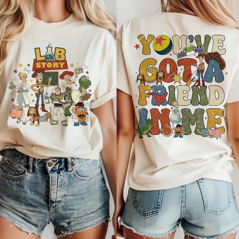 Op de afbeelding: Cr&egrave;mekleurig T-shirt met de tekst "Lab Story" en cartoonfiguren op de voorkant, en "You've Got a Friend in Me" op de achterkant. Het ontwerp toont kleurrijke animatiefiguren in een speelse stijl.