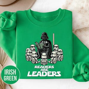 K&ouml;nnte beinhalten: Irisch gr&uuml;nes Sweatshirt mit einer Cartoon-Illustration von Darth Vader, der Stormtroopern vorliest. Der Text "READERS ARE LEADERS" befindet sich darunter. Ein rotes Lichtschwert ist ebenfalls zu sehen.