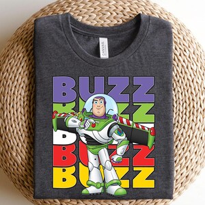 Puede incluir: Camiseta gris oscuro con un gr&aacute;fico de Buzz Lightyear de Toy Story. El dise&ntilde;o incluye la palabra "BUZZ" en letras apiladas y coloridas. Buzz est&aacute; representado con su traje espacial verde y blanco.