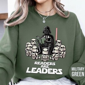 K&ouml;nnte beinhalten: Milit&auml;rgr&uuml;nes Sweatshirt mit einer Grafik von Darth Vader, der Stormtroopern vorliest. Der Text "Readers are Leaders" befindet sich darunter. Ein rotes Lichtschwert ist sichtbar. Das Sweatshirt ist einfarbig.