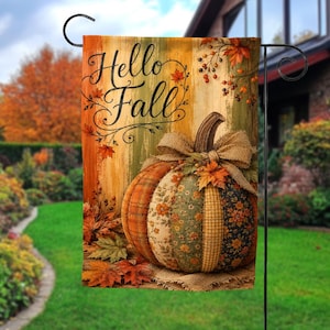 May include: A rectangular garden flag featuring the text Hello Fall in black script. The design displays a large pumpkin made of various fabric patches in orange, green, and floral patterns, topped with a burlap bow and surrounded by autumn leaves.