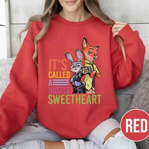 Puede incluir: Una sudadera roja con los personajes Judy Hopps y Nick Wilde de Zootopia. El texto "IT'S CALLED A HUSTLE SWEETHEART" est&aacute; impreso en varios colores. El dise&ntilde;o presenta a los personajes en un estilo de dibujos animados.