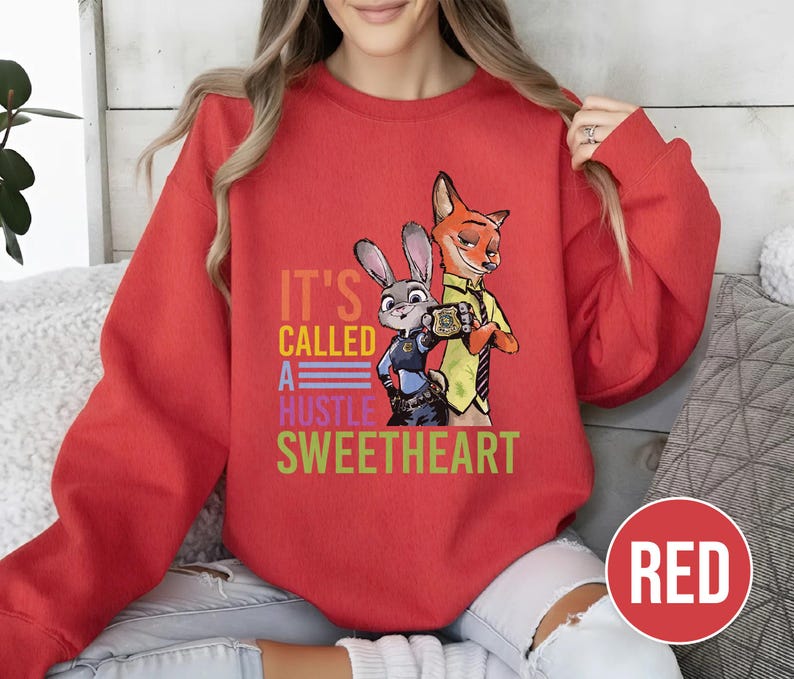Puede incluir: Una sudadera roja con los personajes Judy Hopps y Nick Wilde de Zootopia. El texto "IT'S CALLED A HUSTLE SWEETHEART" est&aacute; impreso en varios colores. El dise&ntilde;o presenta a los personajes en un estilo de dibujos animados.