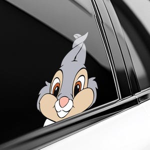 May include: A gray and tan cartoon rabbit sticker, featuring large brown eyes and a pink nose. The rabbit is peeking out from behind a car window, with a white shirt collar visible. The sticker is designed to be applied to a car window.