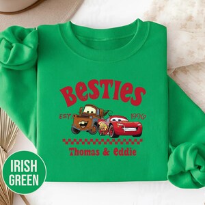 May include: Irish green sweatshirt featuring the text "Besties" in red, along with the year "1996". The design includes cartoon images of two cars, with the names "Thomas & Eddie" below. The sweatshirt has a crew neck and long sleeves.