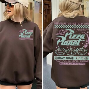 May include: A brown sweatshirt with the Pizza Planet logo on the front and back. The front logo is a stylized design with the words "Pizza Planet" in a retro font. The back features the text "Interstellar Fast-Food Fueling Station" and "Friday Night Kid Night".