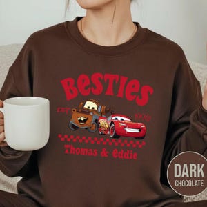 May include: Dark chocolate brown sweatshirt featuring a graphic of two cartoon cars with the text "Besties" in red. The design includes the text "EST 1996" and the names "Thomas & Eddie" in red.